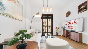 A modern living room with a sofa, a coffee table, a bonsai tree, a TV, and a 'BOOK ME' sign. - Free Housekeeping Visits During Your Stay - Stayshort - Cozy Townsquare 1 Bed Apartment With Balcony That Sleeps 4 (Dubai)