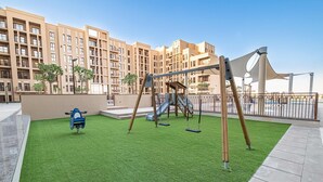 A playground with swings and a slide, surrounded by a green lawn and a building in the background. - Free Housekeeping Visits During Your Stay - Stayshort - Cozy Townsquare 1 Bed Apartment With Balcony That Sleeps 4 (Dubai)