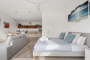 A modern bedroom with a bed, a sofa, a dining area, and a kitchen in the background.