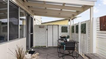 A covered patio with a table and chairs, a grill, and a white fence with greenery.