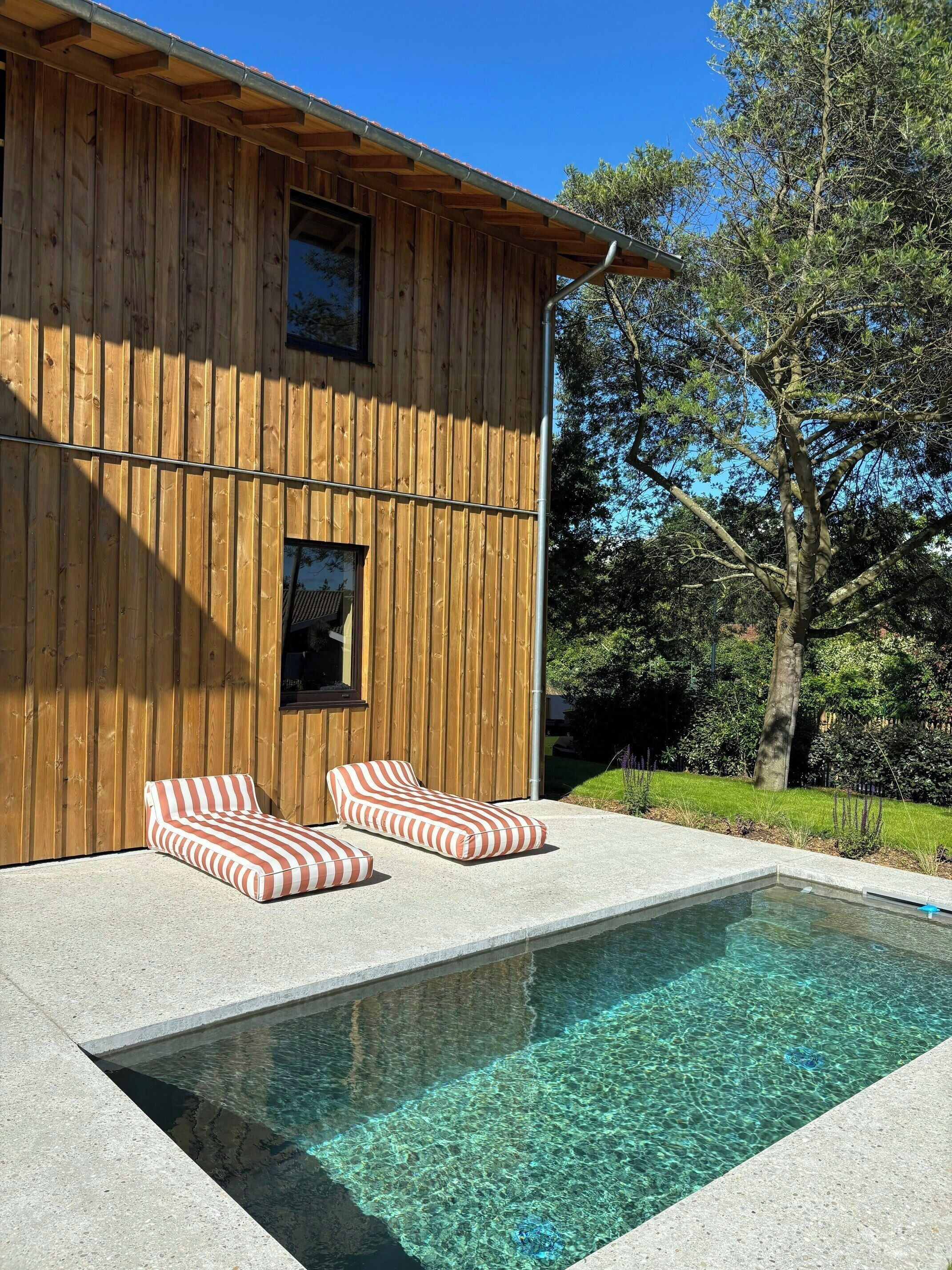 A wooden building with a swimming pool, two striped lounge chairs, and a clear blue sky.