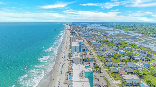 Vista aérea de un pueblo costero con playa, zonas residenciales y un muelle que se adentra en el mar.