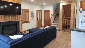 A modern living room with a large dark blue sectional sofa, a fireplace, wooden paneling, a flat-screen TV, and a staircase leading to a kitchen area. - Family Hot Tub, Mountainscape Chalet! Paradise! (Breckenridge)