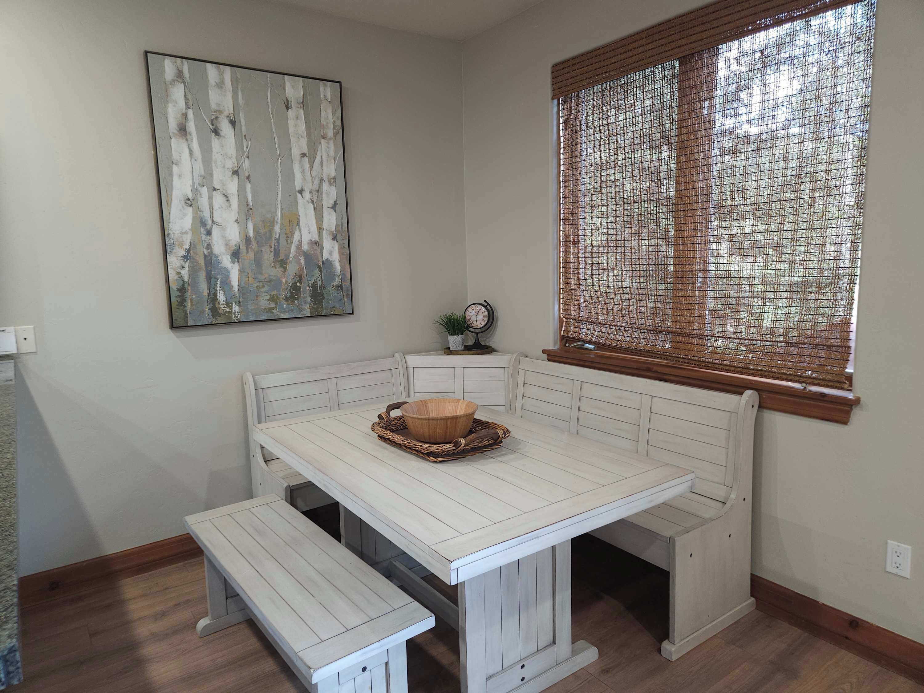 A corner dining area with a white wooden bench and table, a woven basket, and a painting on the wall.
