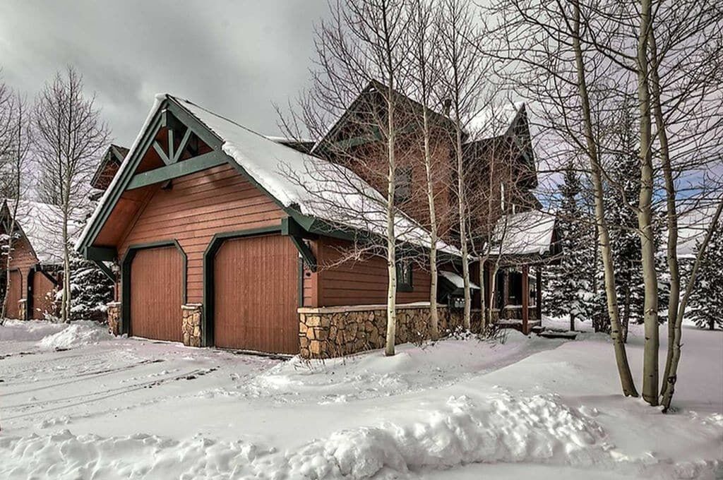 A wooden cabin with a snow-covered roof, stone foundation, and a garage.