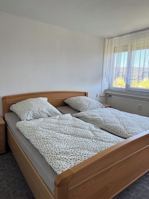 A single bed with a floral bedspread, a wooden headboard, a nightstand, a window with sheer curtains, and a radiator.