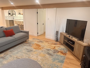 Een woonkamer met een bank, een televisie, een vloerkleed en een deur naar een keuken.