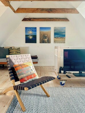 A loft-style room with a high ceiling, wooden beams, a woven chair, a flat-screen TV, and three landscape paintings on the wall. - Townhouse from 1862 with sunsets in Thorøhuse (Assens)