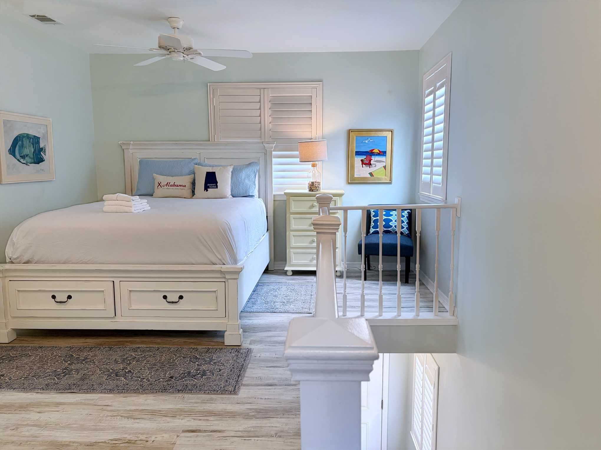 A bedroom with a bed, bedside tables, a nightstand, a crib, a ceiling fan, and artwork on the walls.