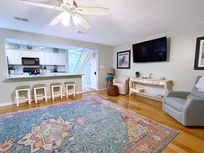 A living room with a large rug, a grey sofa, a TV, and a kitchen area with white cabinets and a microwave. - Bayview 66 Townhome | Orange Beach Condo with Deeded Boat Slip + Boat Launch | Wolf Bay (Orange Beach)