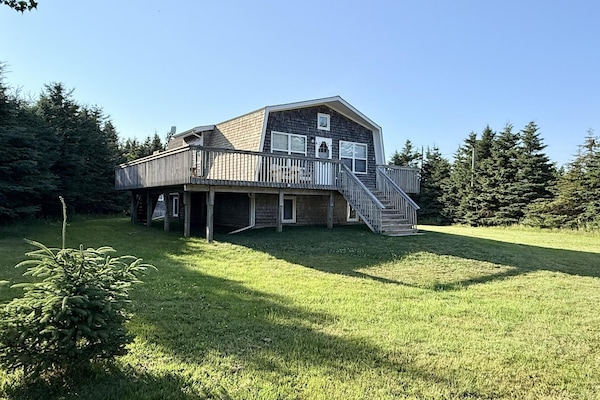 Raised wooden deck wraps around the cedar-shingled cottage with a gambrel roof, surrounded by green lawn and evergreens.