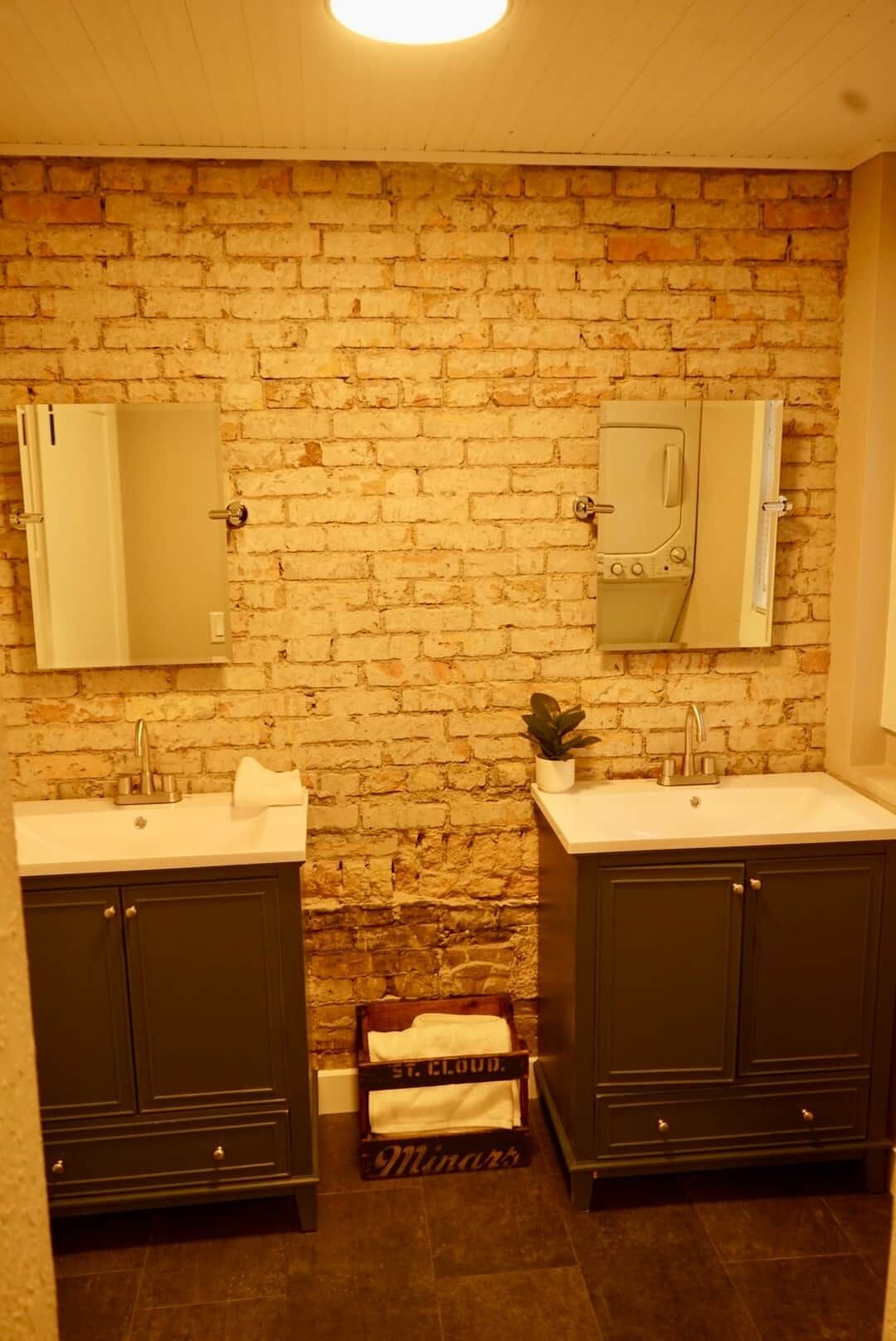 A bathroom with a brick wall, two sinks, dark cabinets, and a small plant.
