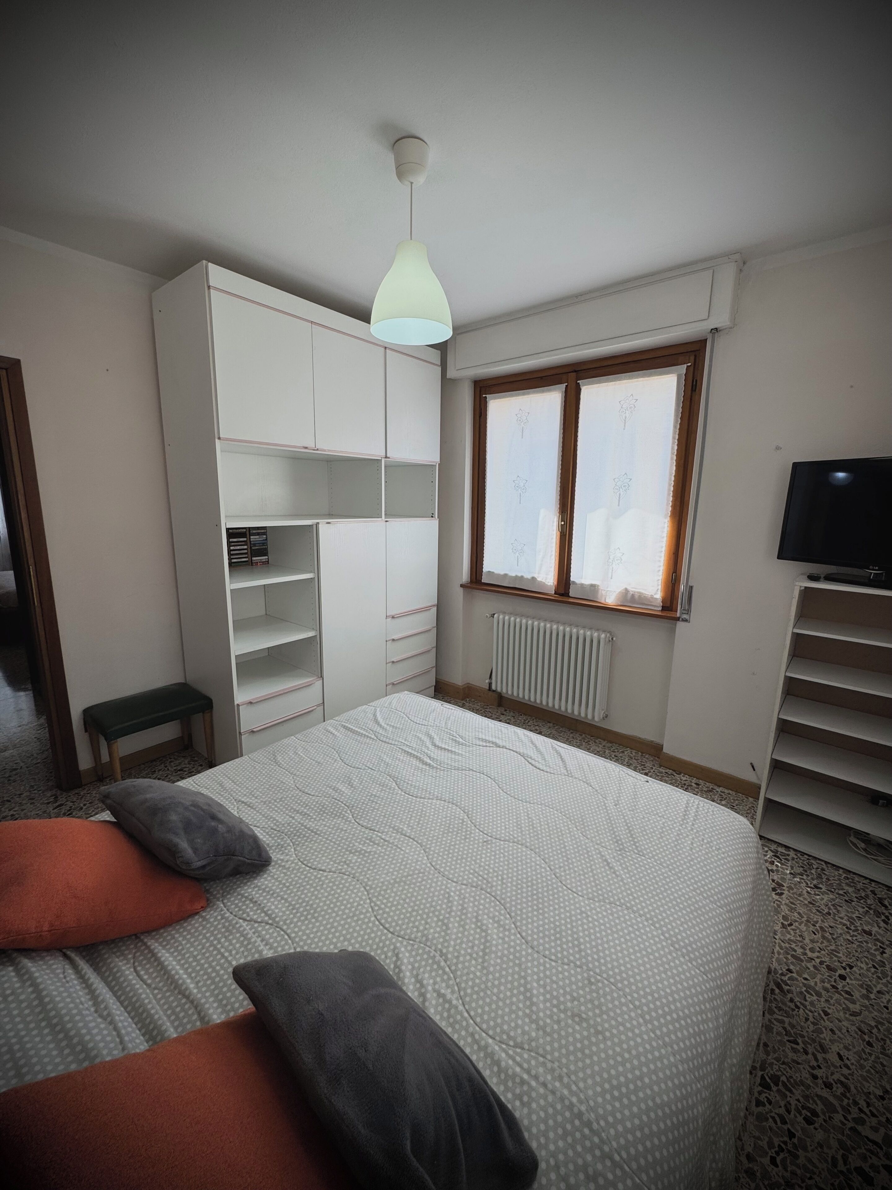 A bedroom with a bed, a wardrobe, a radiator, a window with curtains, and a television.