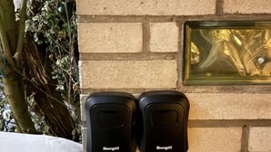Twee zwarte intercoms bevestigd aan een bakstenen muur, met een raam en een plant op de achtergrond.