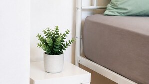 Een klein nachtkastje, wit van kleur, met een potplantje, een bed met een groen kussen en een wit hoofdbord.