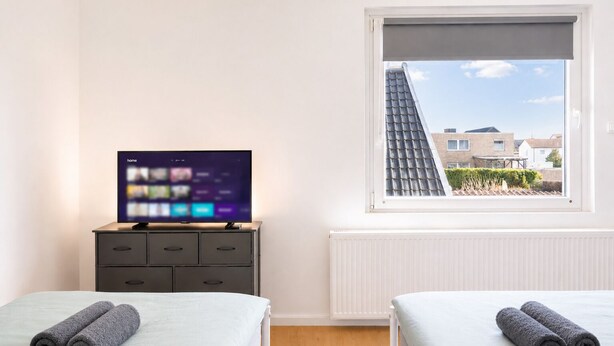 Een slaapkamer met een televisie op een dressoir, twee bedden met handdoeken, een raam met uitzicht op gebouwen en een radiator.