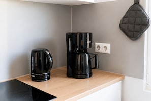 A kitchenette with a coffee maker and a kettle on a wooden countertop, a wall socket, and a hanging pot rack.