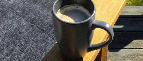 Une tasse en métal posée sur un banc en bois, avec en arrière-plan une prairie verdoyante et une étendue d’eau.