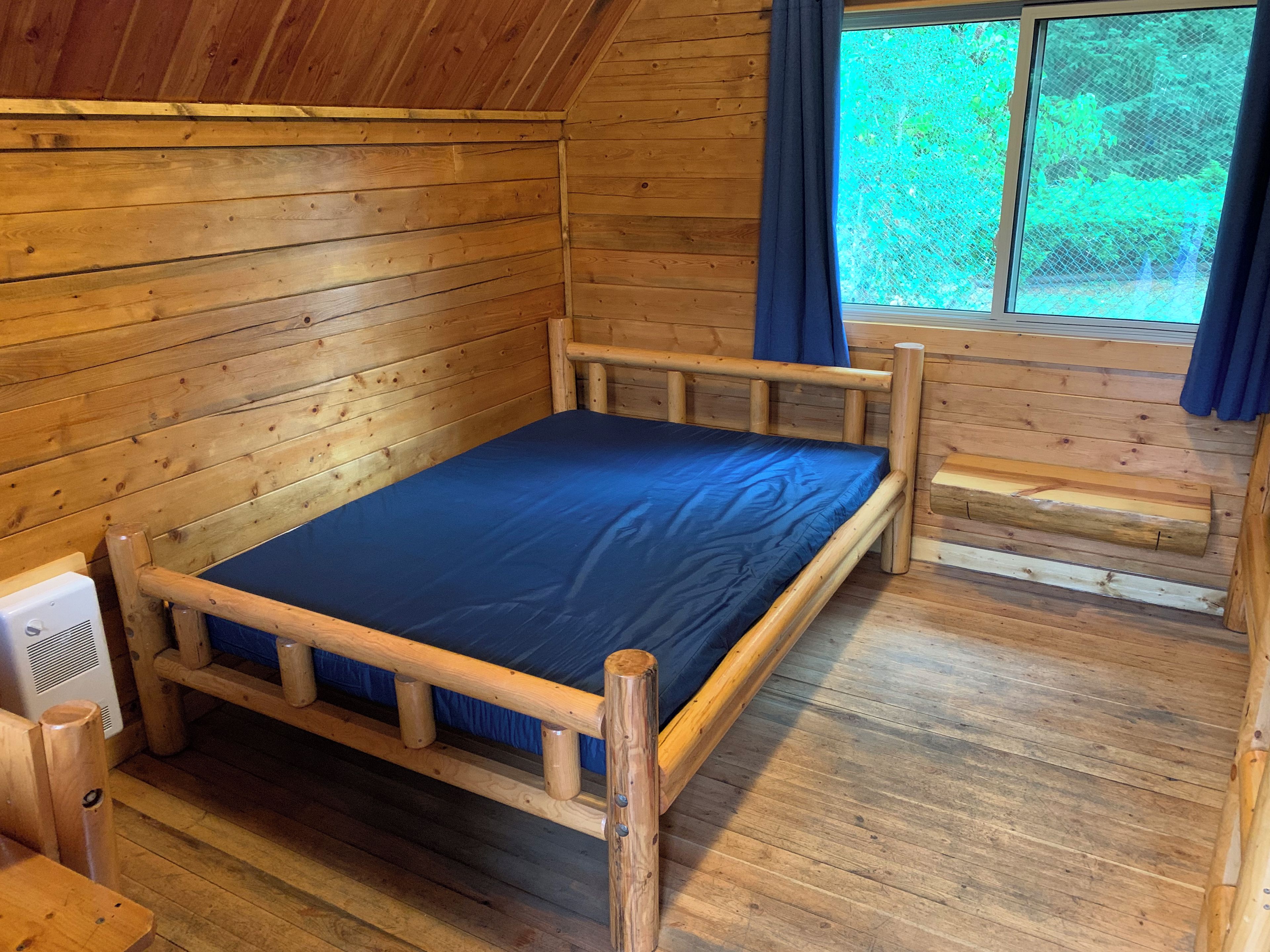 A wooden cabin room with a single bed, a small table, and a window with curtains.
