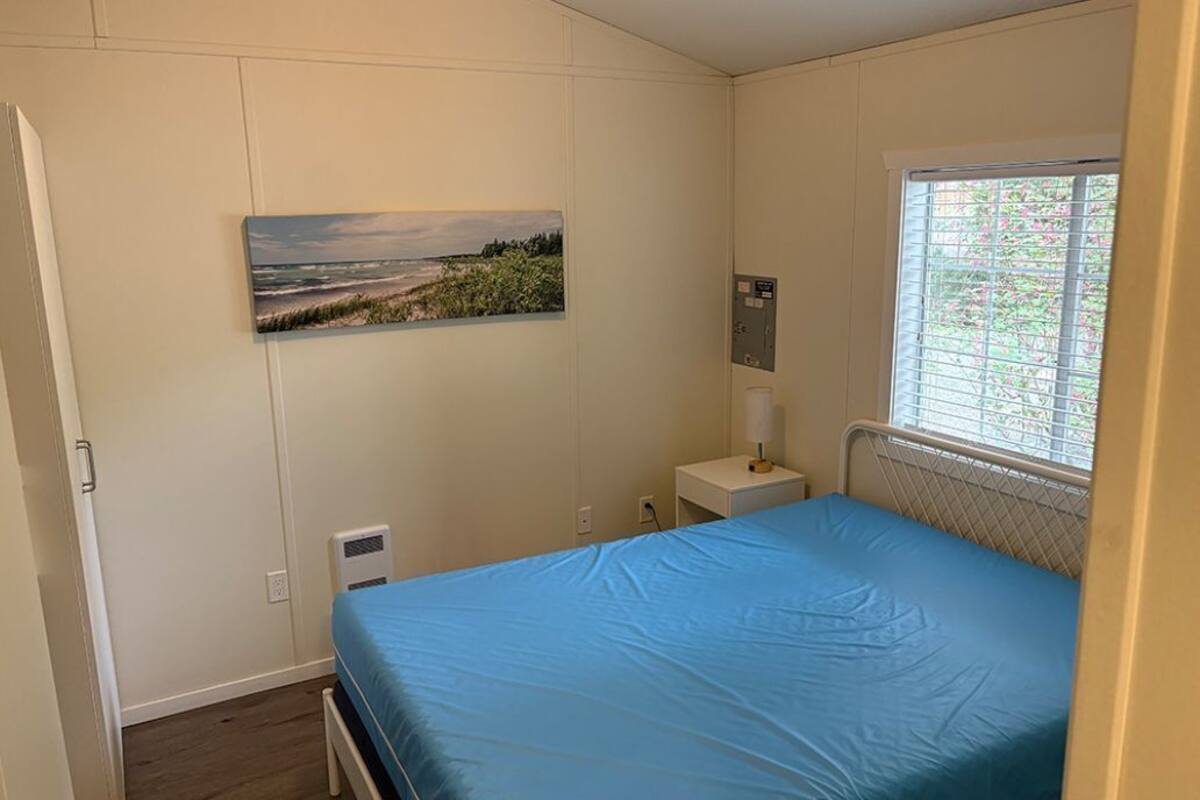 A bedroom with a bed, a window with blinds, a bedside table with a lamp, and a wall-mounted electrical outlet.