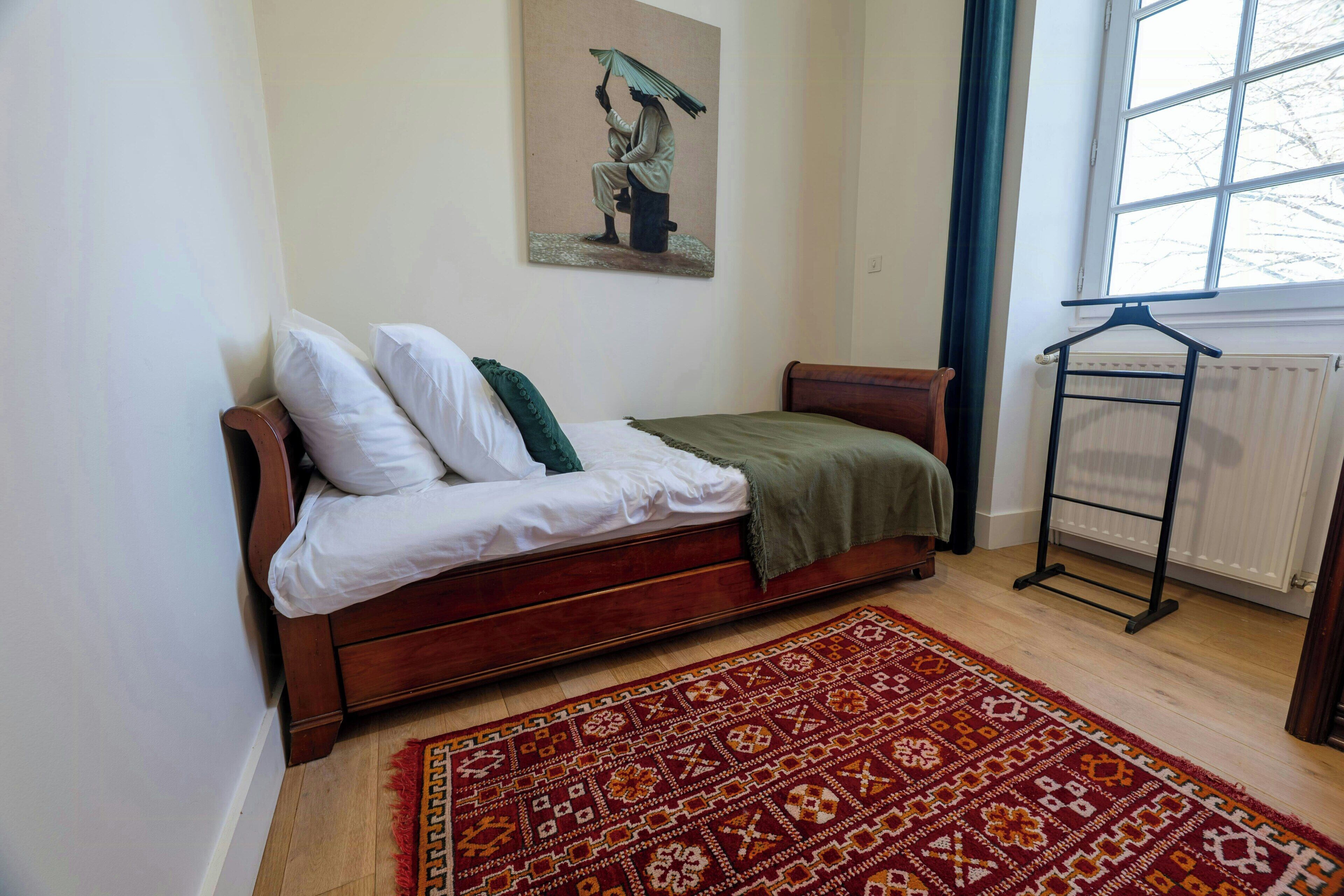 Un dormitorio con una cama de madera, un tapete estampado, un radiador y una ventana con vista a árboles.