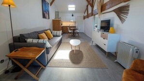 Un salon avec un canapé, une table basse et un téléviseur. Au fond, on aperçoit une cuisine avec une table à manger et des chaises.