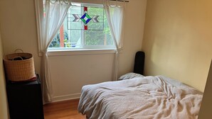 Una camera da letto con un letto, una finestra con tende, un cesto su un tavolo e un altoparlante a parete.