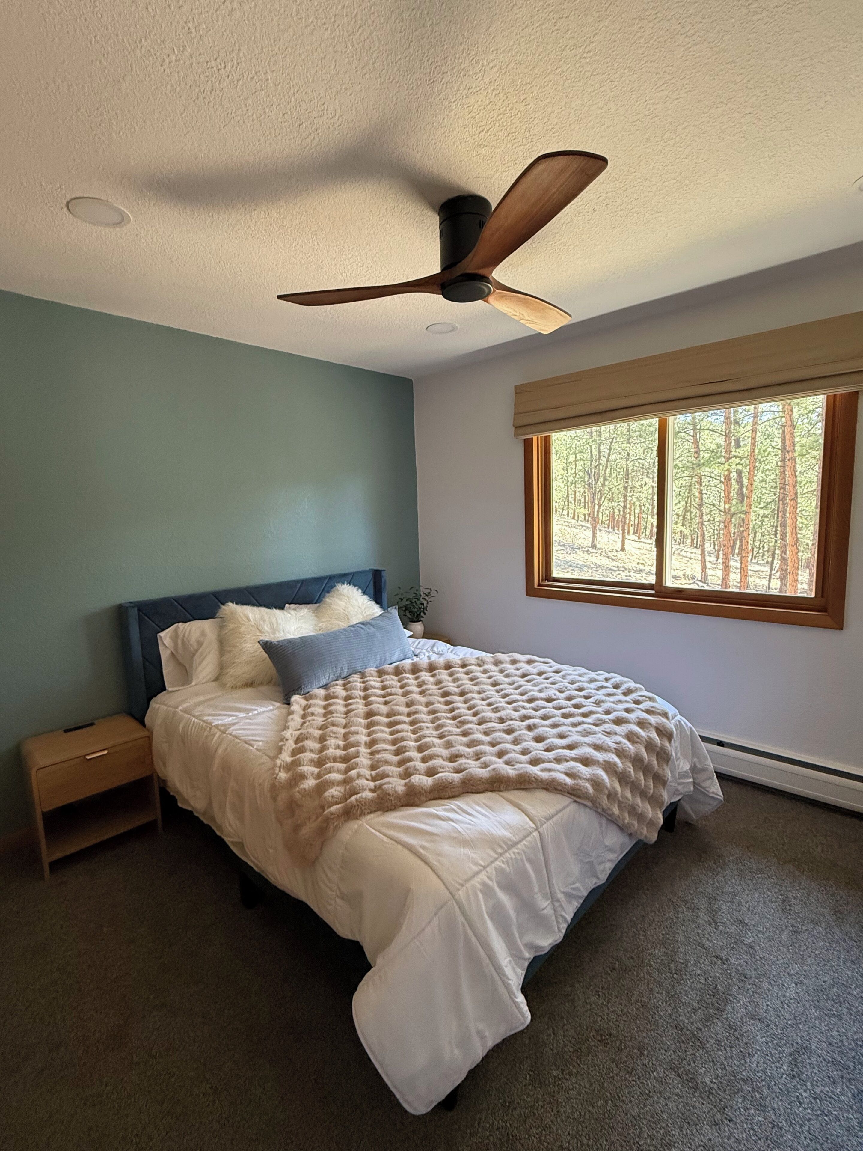 A bedroom with a bed, a ceiling fan, a window with a view of trees, and a nightstand.