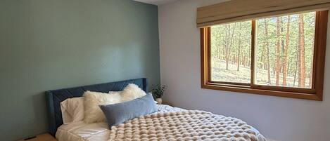 A bedroom with a bed, a ceiling fan, a window with a view of trees, and a nightstand.