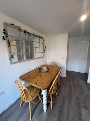 Een eetgedeelte met een houten tafel, vier stoelen, een grote spiegel met decoratieve lijst, en een witte deur op de achtergrond.