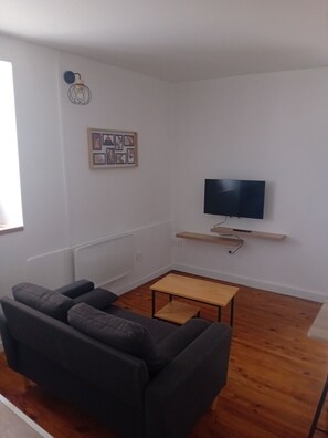 Ein Wohnzimmer mit einer Couch, einem Fernseher auf einem Holzregal und einem gerahmten Bild an der Wand.