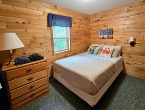 Quarto com painéis de madeira, uma cama, cômoda, janela com persianas e luminária fixada na parede.