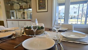 Un tavolo da pranzo apparecchiato con piatti, bicchieri e posate, una ciotola decorativa con piante verdi e vista su una casa attraverso una finestra.