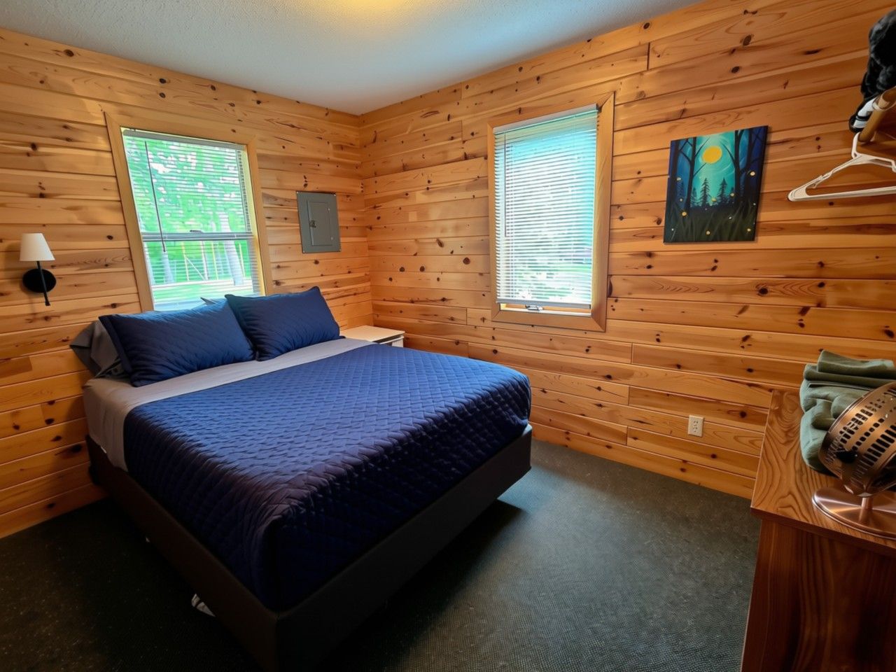 Quarto com paredes de madeira, uma cama com roupa de cama azul, duas janelas com persianas, um quadro na parede e um aquecedor sobre uma mesa de madeira.