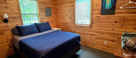 Quarto com paredes de madeira, uma cama com roupa de cama azul, duas janelas com persianas, um quadro na parede e um aquecedor sobre uma mesa de madeira.