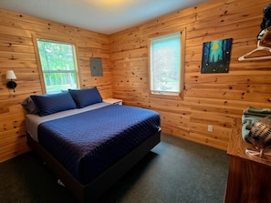 A room with wooden walls, a bed with blue bedding, two windows with blinds, a painting on the wall, and a heater on a wooden table. - Tamarack at Evergreen Bay Resort on Boot Lake (Park Rapids)