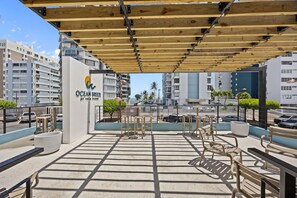 A rooftop terrace with outdoor seating, a sign for Ocean Breeze at Asia Term, and modern buildings in the background. - Isla Verde Ocean Breeze | Luxury Beach Suites 2E (Carolina)