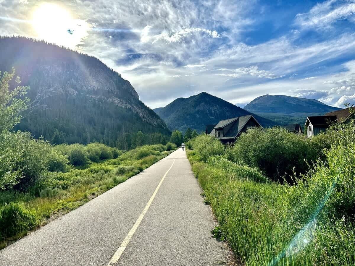 A road flanked by lush greenery and houses, leading towards distant mountains under a partly cloudy sky.
