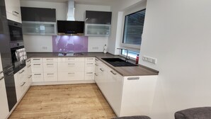 Una cocina moderna con armarios blancos, horno empotrado, microondas, televisor y ventana con persianas.
