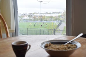 Ein Zimmer mit großem Fenster mit Blick auf ein Sportfeld, einem Esstisch mit Schüssel und einer Tasse sowie einem Stuhl.