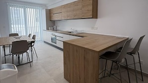 Una cocina moderna con una isla central, armarios de madera, un comedor con sillas y un ventanal con persianas.