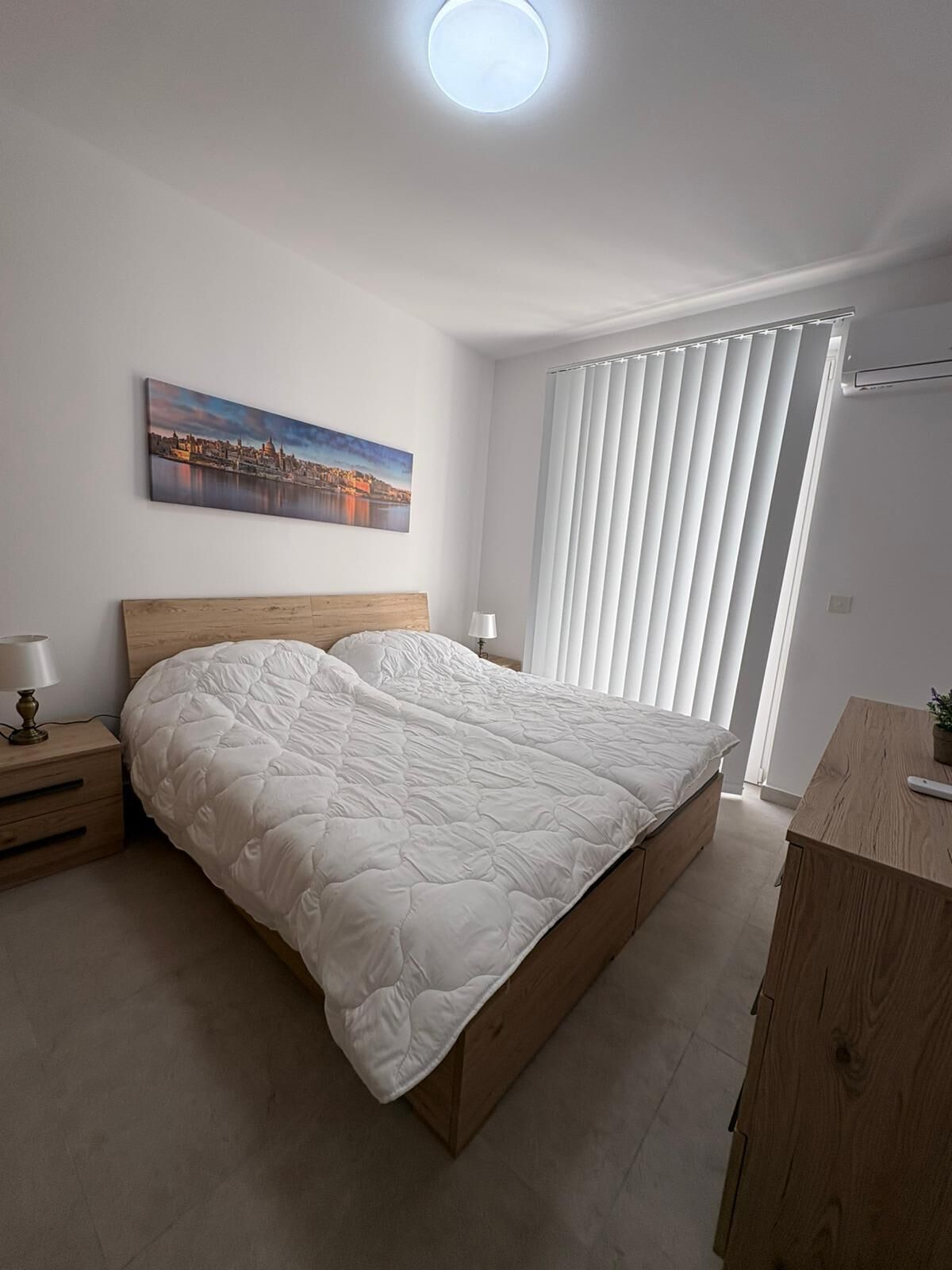 A bedroom with a bed, a nightstand, a lamp, a painting, a window with blinds, and a desk with a plant.