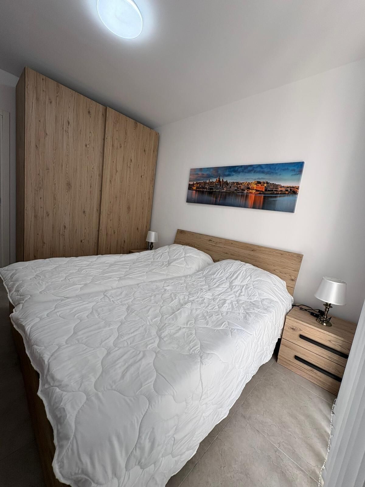A bedroom with a large bed, a wooden wardrobe, a nightstand with a lamp, and a cityscape painting on the wall.