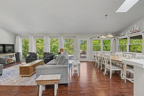 A spacious living room with a sofa, coffee table, and TV, a dining area with chairs and a table, and large windows with a view of greenery. - Beaver Lake Access Retreat w Deck & Grill! (Garfield)