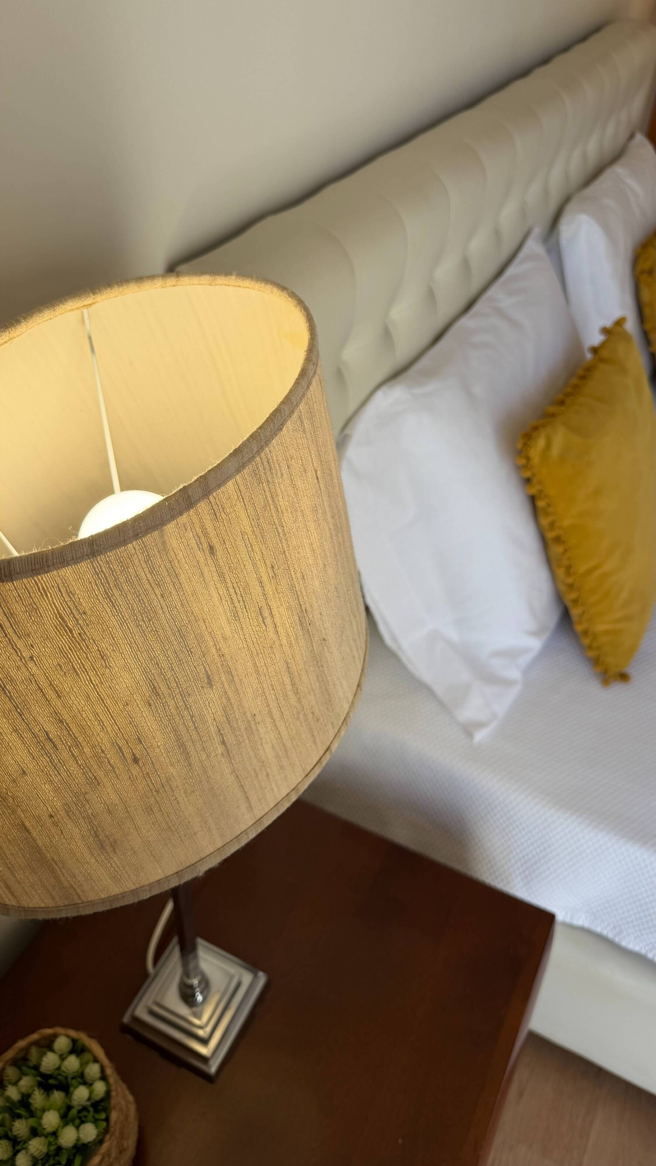 A lamp with a textured shade on a wooden table, a white sofa with a tufted backrest, and a yellow pillow.