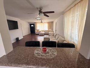 Ein Wohnzimmer mit einem Ecksofa, einem Esstisch mit Stühlen, einem Deckenventilator und einem an der Wand befestigten Fernseher.