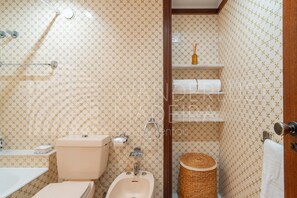A bathroom with patterned tiles, a toilet, bidet, sink, and a shelf with toiletries. - Baía Seaview | 2-Bedroom Apartment (Funchal)