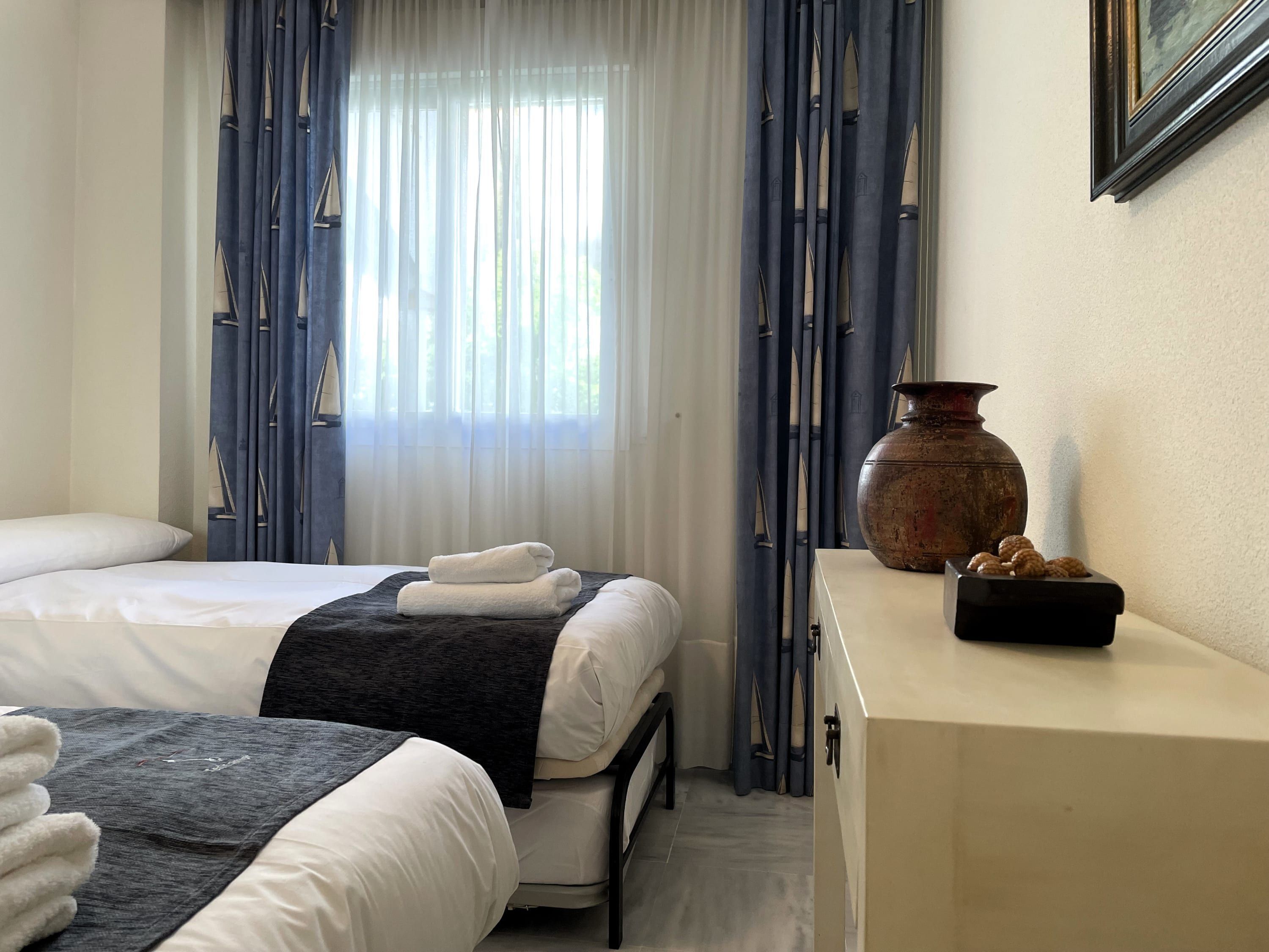 A hotel room with two beds, a large window with sheer curtains, a bedside table with a vase, and a wall-mounted picture.