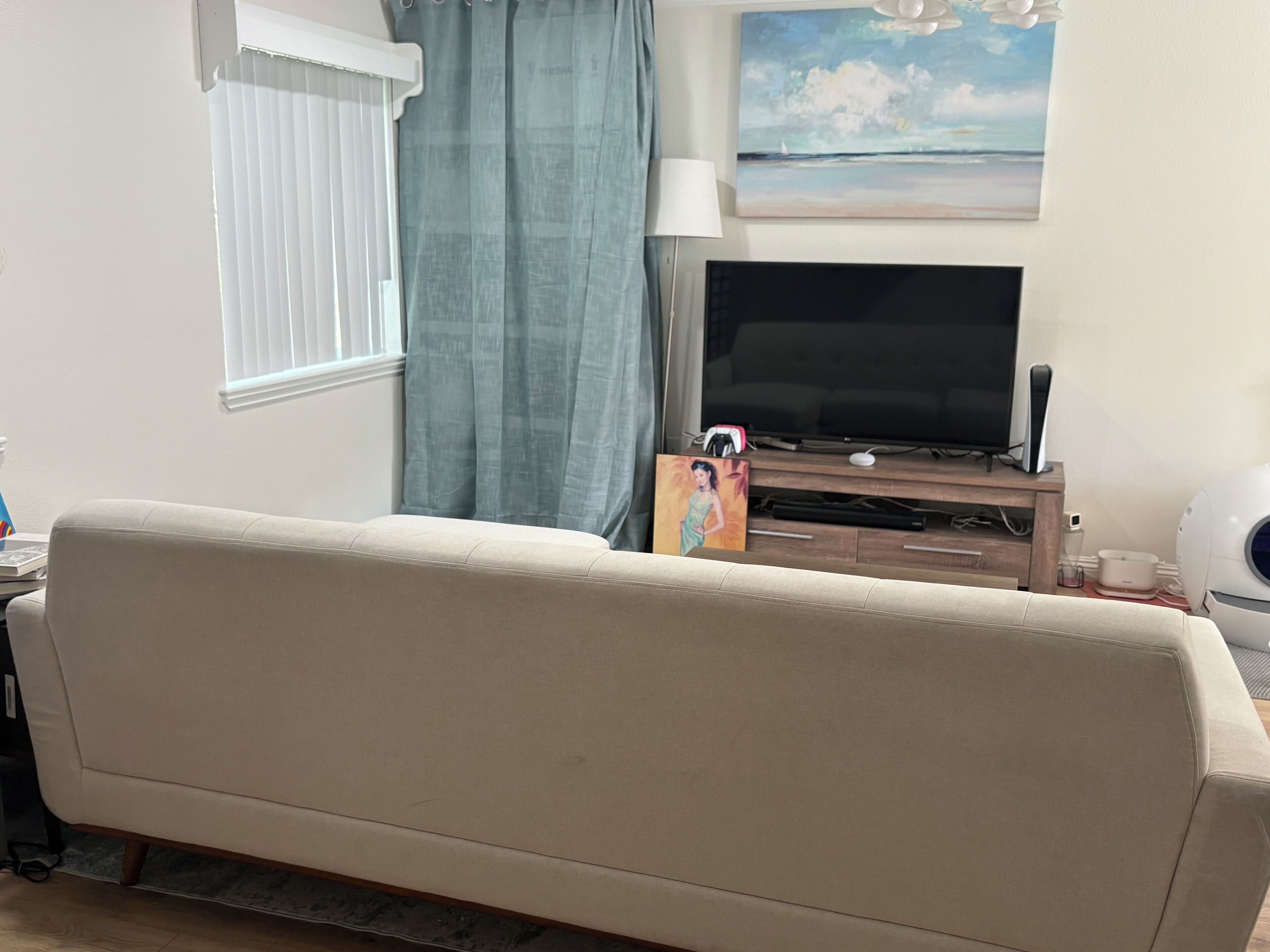 Ein Wohnzimmer mit einem beigefarbenen Sofa, einem Flachbildfernseher, einem Gemälde und einem Fenster mit Jalousien.