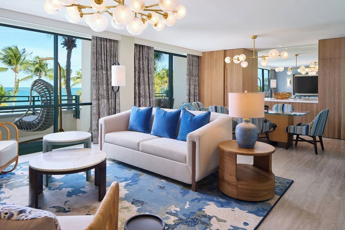A modern hotel room with a sofa, round tables, a dining area, and a large window with a view of palm trees and the ocean.